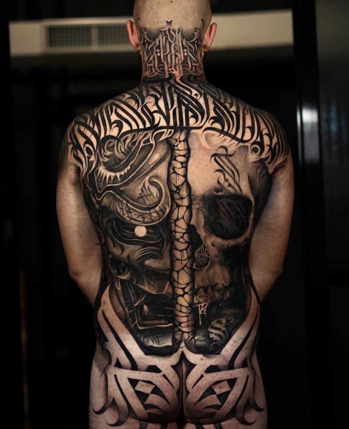 Full back tattoo featuring dual imagery with detailed skull and biomechanical elements connected by spinal design and tribal pattern at the lower back